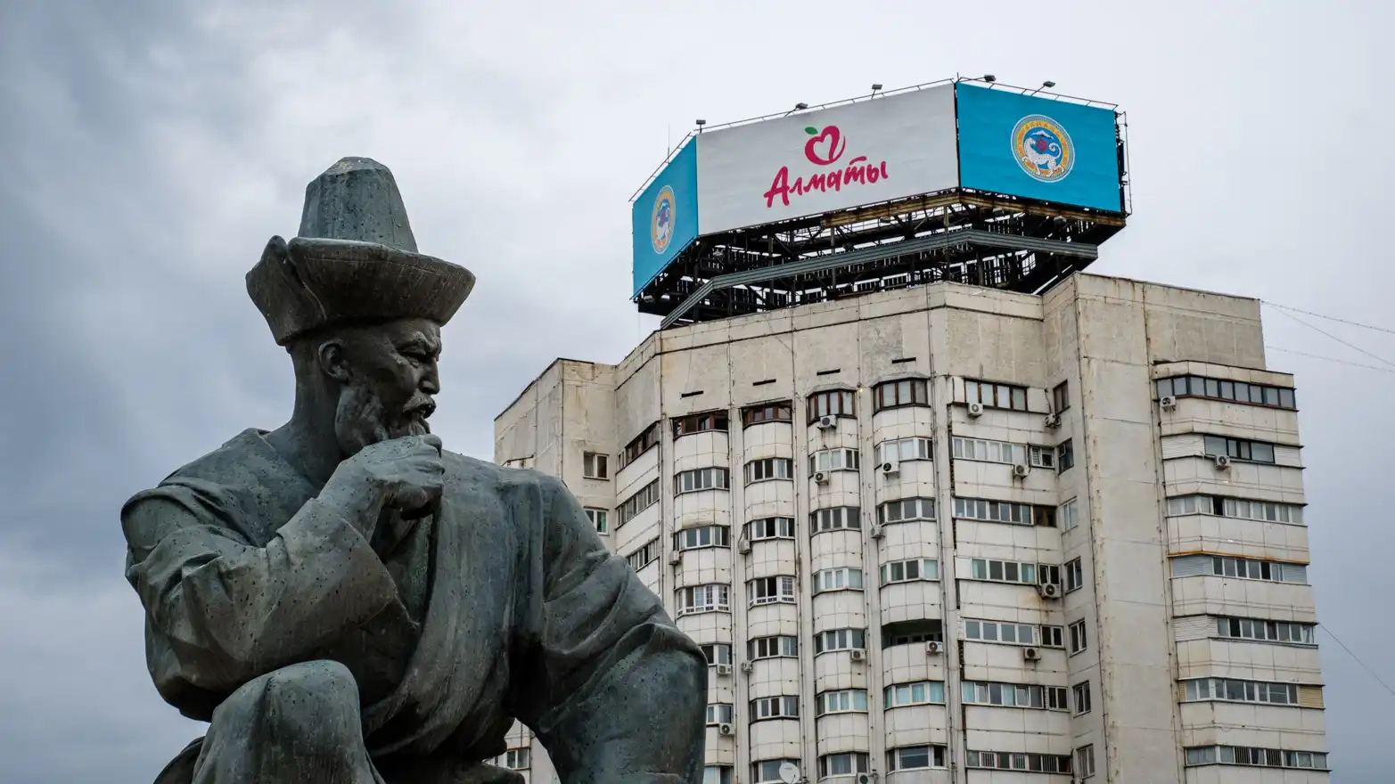 Socha s vysokým panelákem v pozadí. Na střeše paneláku je nápis ALMATY.